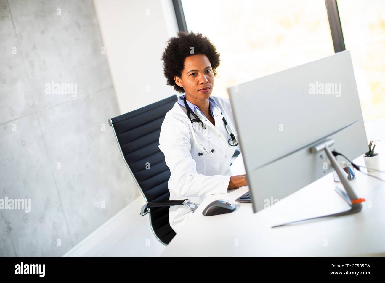 Donna Africana americana medico indossando camice bianco con stetoscopio seduta dietro la scrivania in ufficio Foto Stock