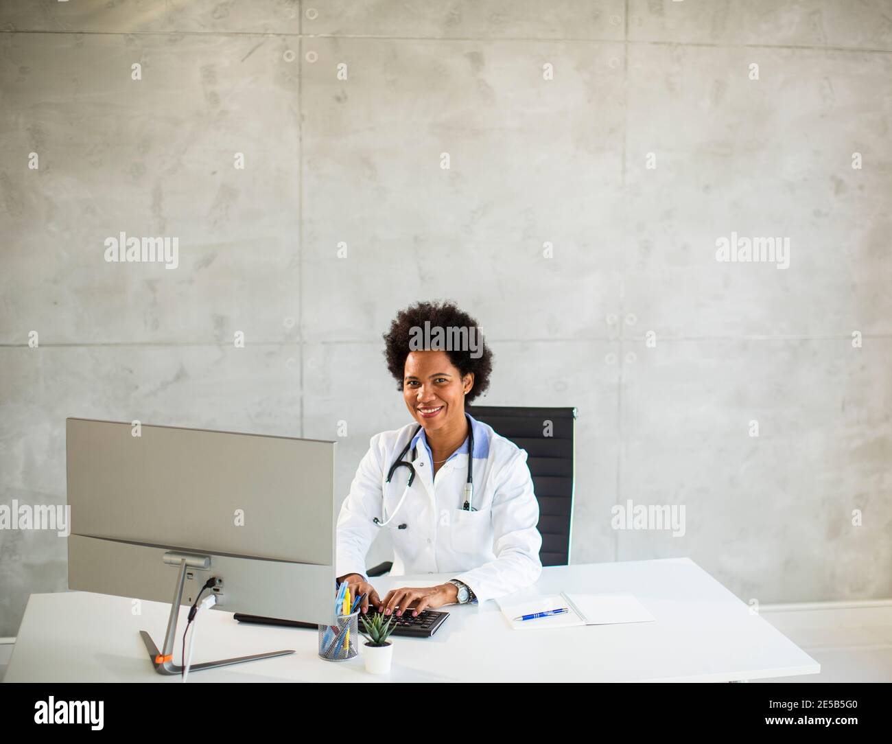 Donna Africana americana medico indossando camice bianco con stetoscopio seduta dietro la scrivania in ufficio Foto Stock