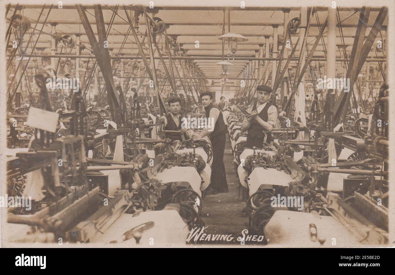 Capannone di tessitura in un mulino di cotone Lancashire, Bury. Tre ragazzi sono al lavoro tra i macchinari del mulino, una donna è sullo sfondo, all'inizio del 20 ° secolo Foto Stock