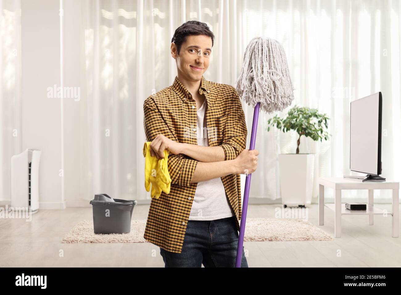 Giovane uomo che tiene un mop di pulizia e un paio di guanti in soggiorno Foto Stock