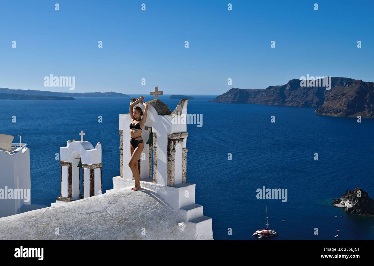 Giovane ragazza in posa sulla cima di una chiesa greco-ortodossa con vista panoramica sul Mar Egeo e la caldera nell'isola di Santorini, Cicladi Grecia. Foto Stock