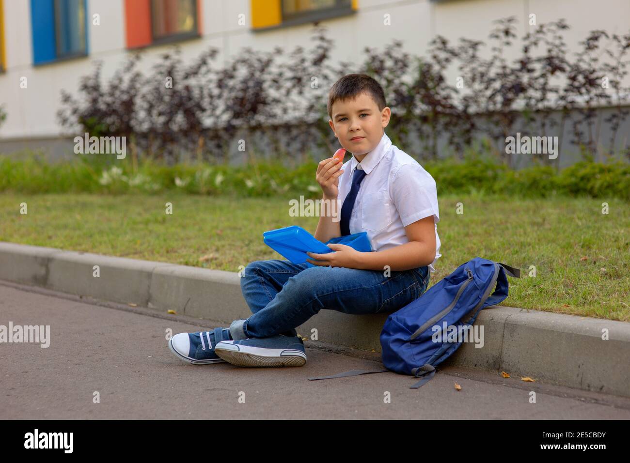 il ragazzo in una camicia bianca con una cravatta blu, tiene una scatola blu per il pranzo e una fetta di mela, guarda la macchina fotografica Foto Stock