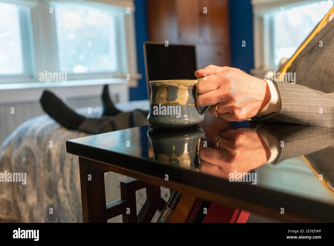 Lavorare da casa l'uomo con il computer portatile si siede sul letto tenendo una tazza di caffè indossando tuta, cravatta e con piedi di calza, umorismo Foto Stock