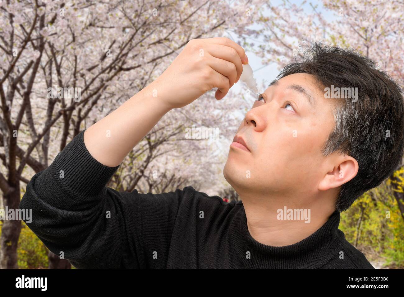 L'uomo asiatico di mezza età che usa gocce di occhio vicino agli alberi in fiore. Concetto di allergia. Foto Stock