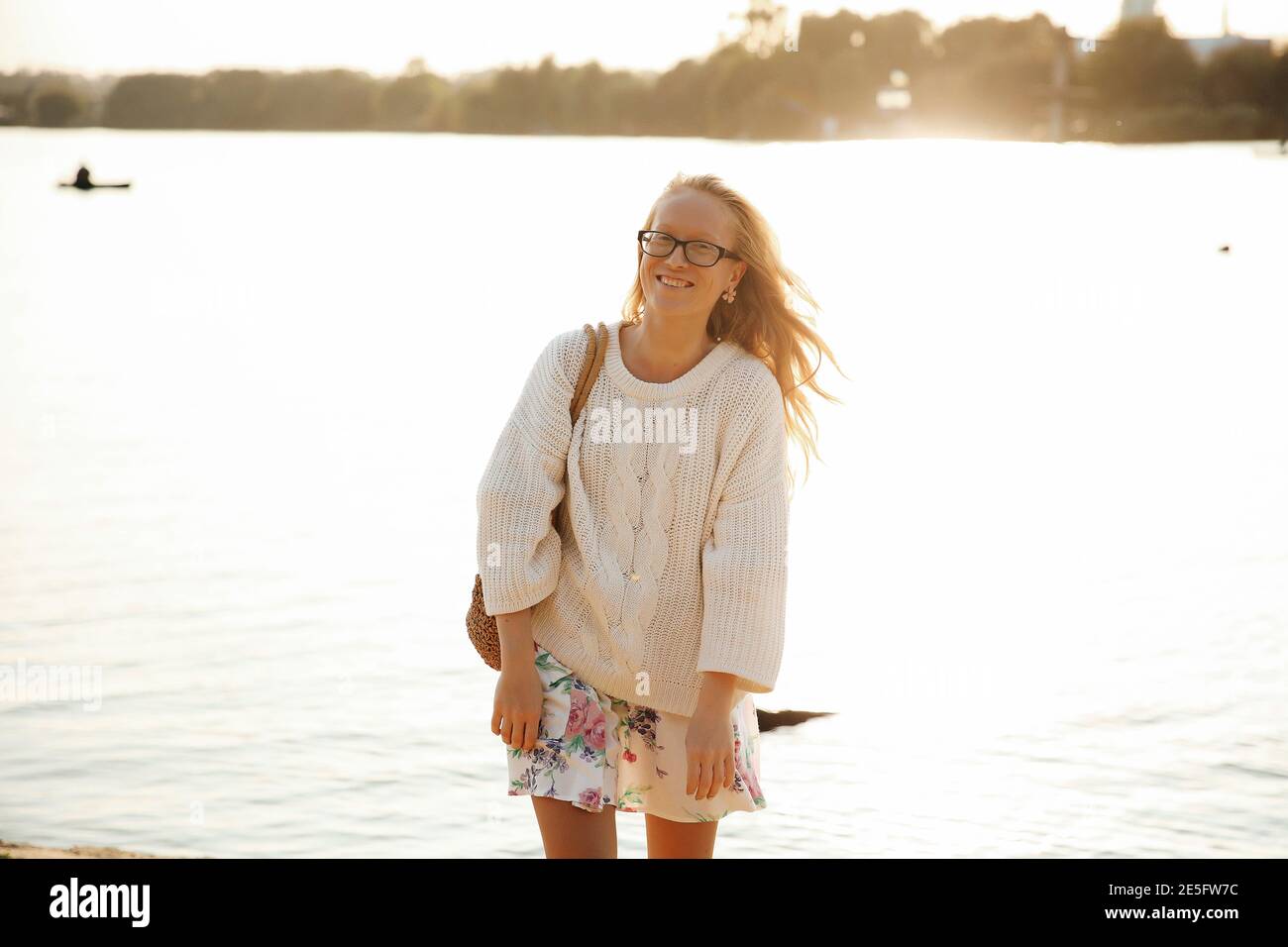 Questa foto mostra una giovane bionda che cammina sulla spiaggia in un vestito e maglione con fiori in una giornata di sole. Foto Stock