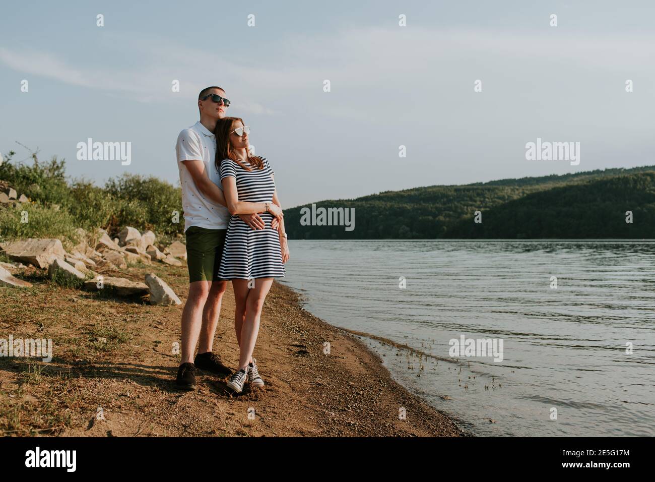 Ritratto di coppia innamorata in piedi sulla spiaggia al tramonto. Tutta la lunghezza di giovane donna e uomo che si agganava sul lago nella calda sera d'estate. Foto Stock