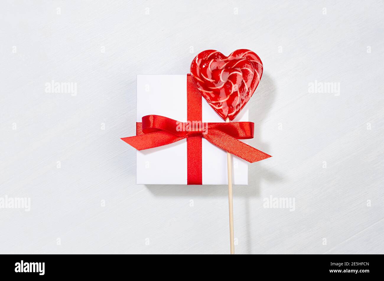 Festive Love background - scatola regalo bianca con nastro di seta rossa e cuore dolce lollipop su tavola di legno bianco, primo piano. Foto Stock