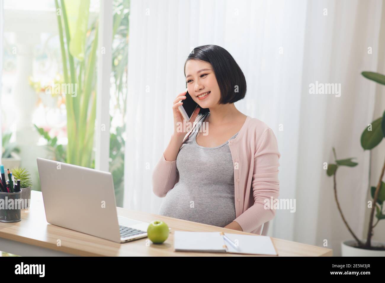 Donna d'affari incinta che lavora con il computer portatile e parla al telefono in un accogliente e luminoso ufficio. Foto Stock