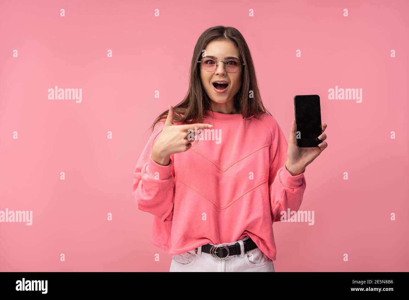 Foto di attraente signora in occhiali tiene smartphone, legge o scrive blog. Indossa casual t-shirt rosa pantaloni bianchi isolato colore rosa sfondo. Foto Stock