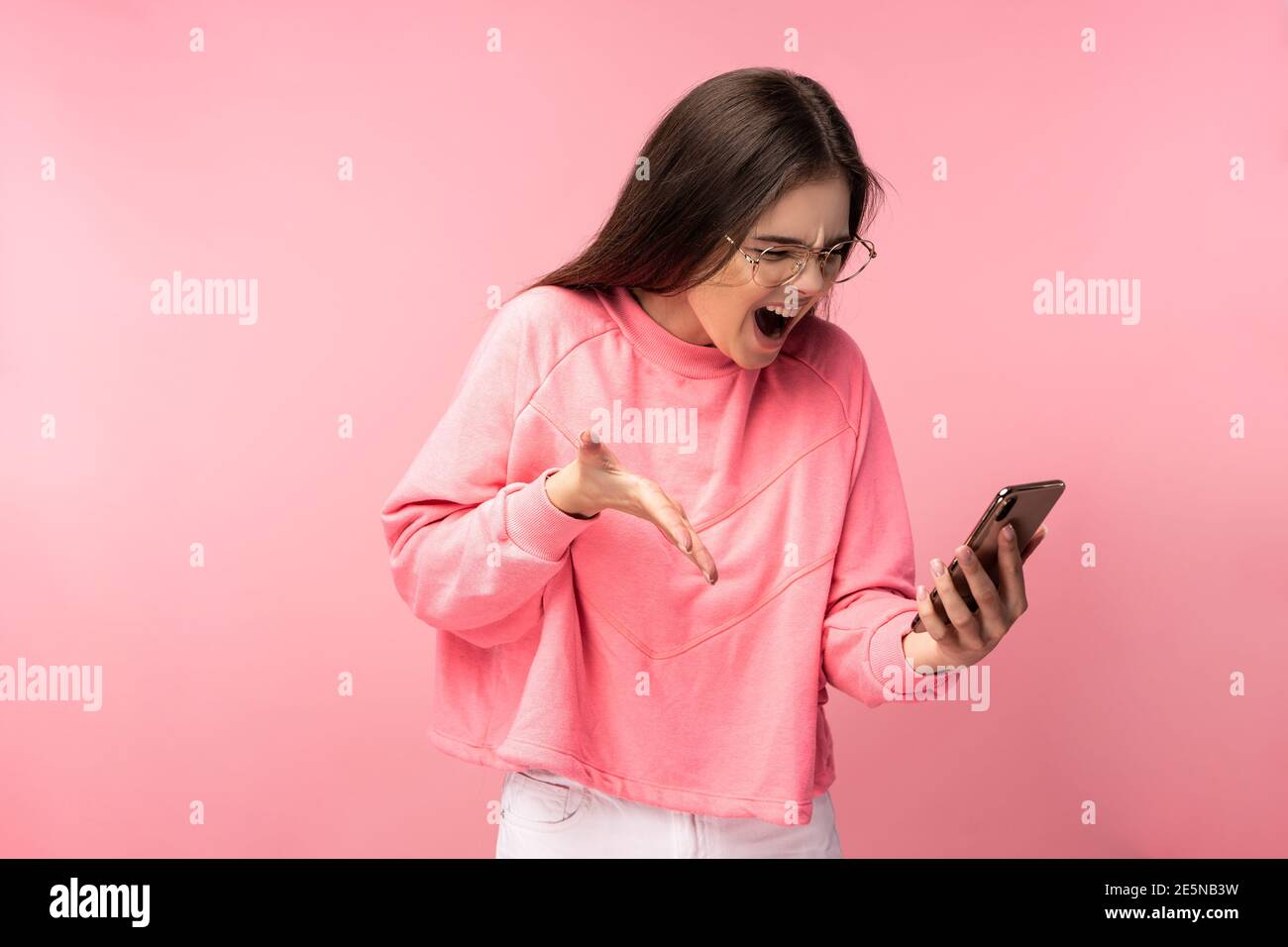 Foto di attraente signora in occhiali tiene il telefono e urla, deluso legge commenti sul blog. Indossa casual t-shirt rosa pantaloni bianchi isolati Foto Stock