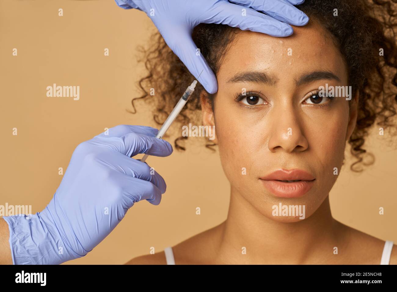 Ritratto di bella giovane donna che guarda la fotocamera mentre ottiene iniezioni viso isolato su sfondo beige. Bellezza, concetto di cosmetologia Foto Stock