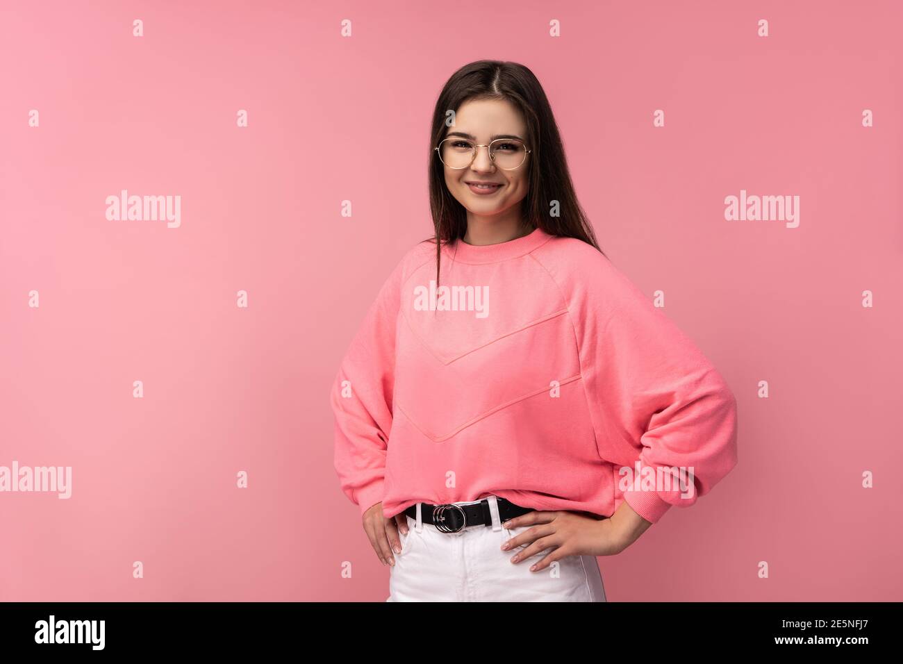 Foto di attraente donna in occhiali sorride e tiene le mani sui fianchi. Indossa casual t-shirt rosa pantaloni bianchi isolato colore rosa sfondo Foto Stock