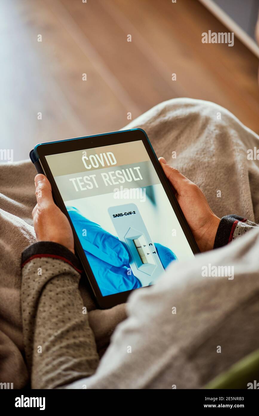 primo piano di un giovane caucasico in pigiama, seduto su un divano avvolto in una coperta grigia circa per verificare il risultato del suo covid test in un'app simulata Foto Stock