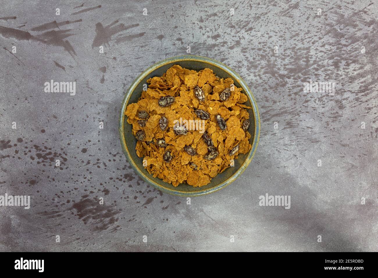 Vista dall'alto di una ciotola piena di fiocchi di crusca e uvetta, cereali per la colazione su sfondo grigio. Foto Stock