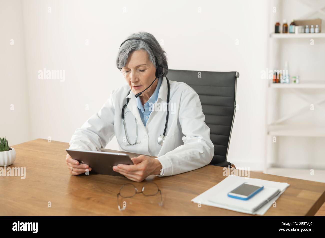Dottore senior in un camice da laboratorio con cuffia seduta alla scrivania, partecipando a una conferenza medica online, leggendo l'elenco prescritto Foto Stock