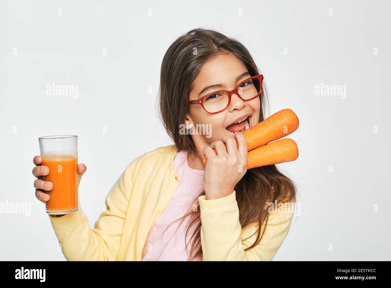 Bambina di razza mista che mangia carote e beve succo di carota, su sfondo grigio. stile di vita sano per i bambini Foto Stock