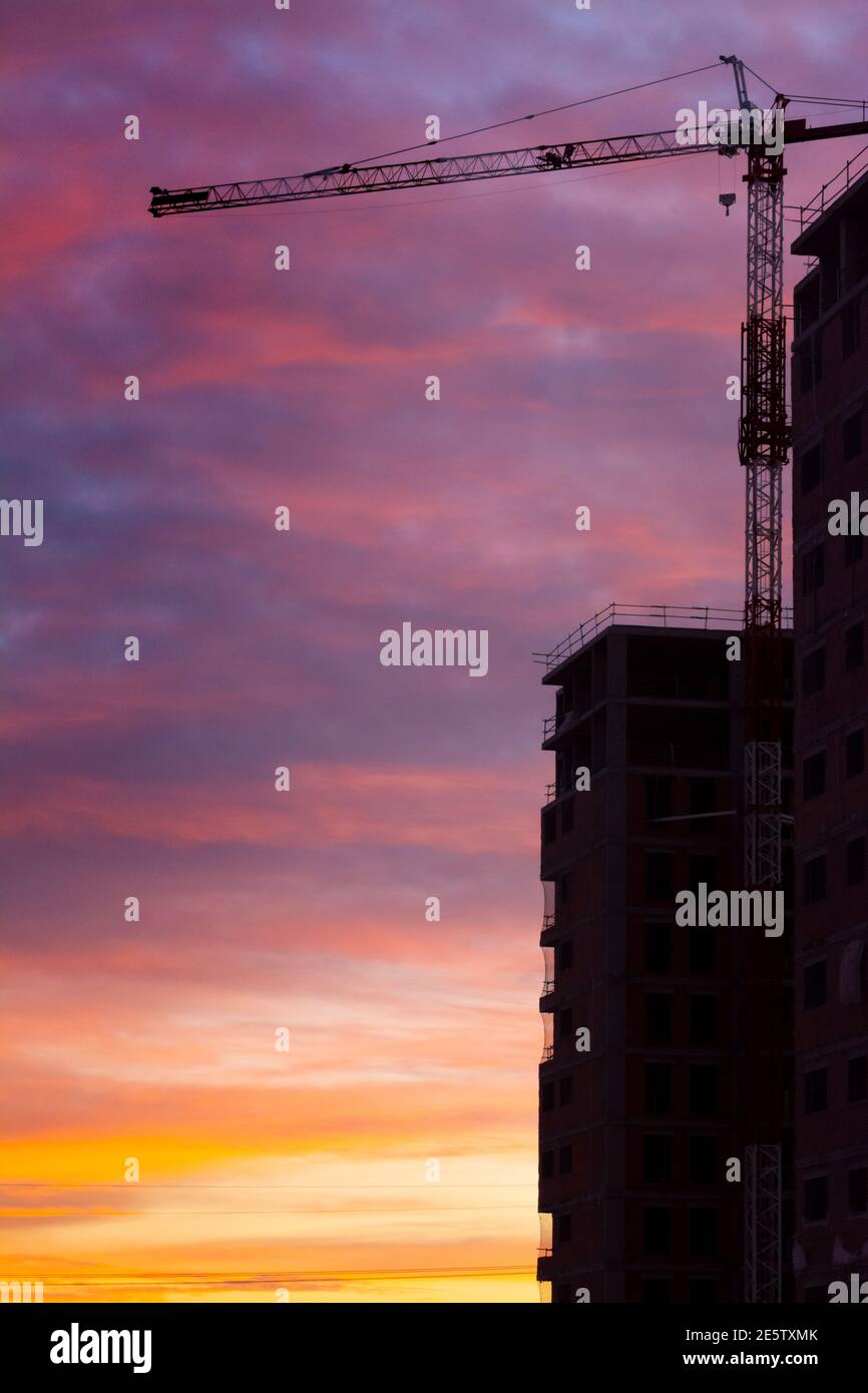 Sagoma di una gru e lato di un edificio in costruzione con cielo blu e giallo e nuvole arancioni al tramonto o all'alba Foto Stock