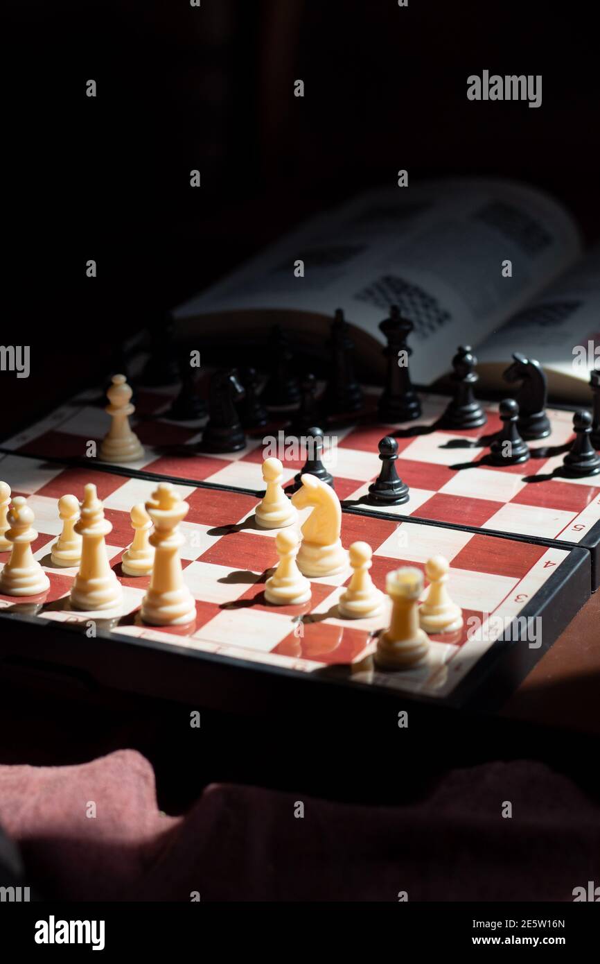 Una scacchiera che mostra l'apertura spagnola e un libro a scacchi aperto su uno sfondo scuro. L'immagine rappresenta una profonda comprensione e studio degli scacchi. Foto Stock
