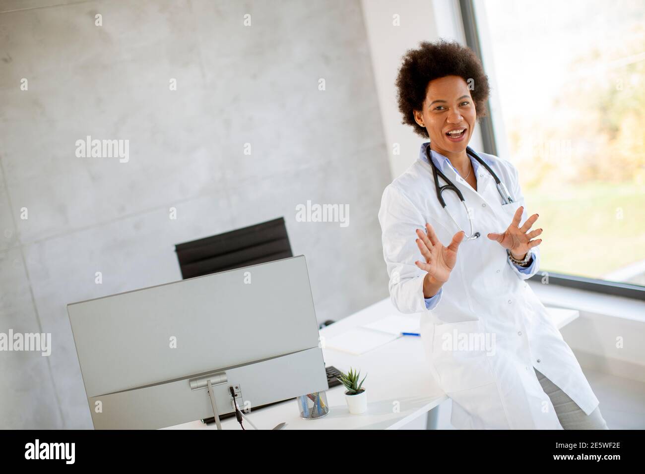 Donna Africana americana medico indossando camice bianco con stetoscopio in piedi dalla scrivania in ufficio Foto Stock