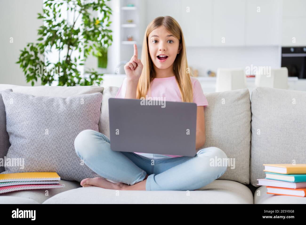 Foto a lunghezza intera di un'intelligente bambina studia a distanza report di pensiero del notebook ottieni un'idea incredibile alza l'indice con il dito verso l'alto sedete le gambe del divano Foto Stock