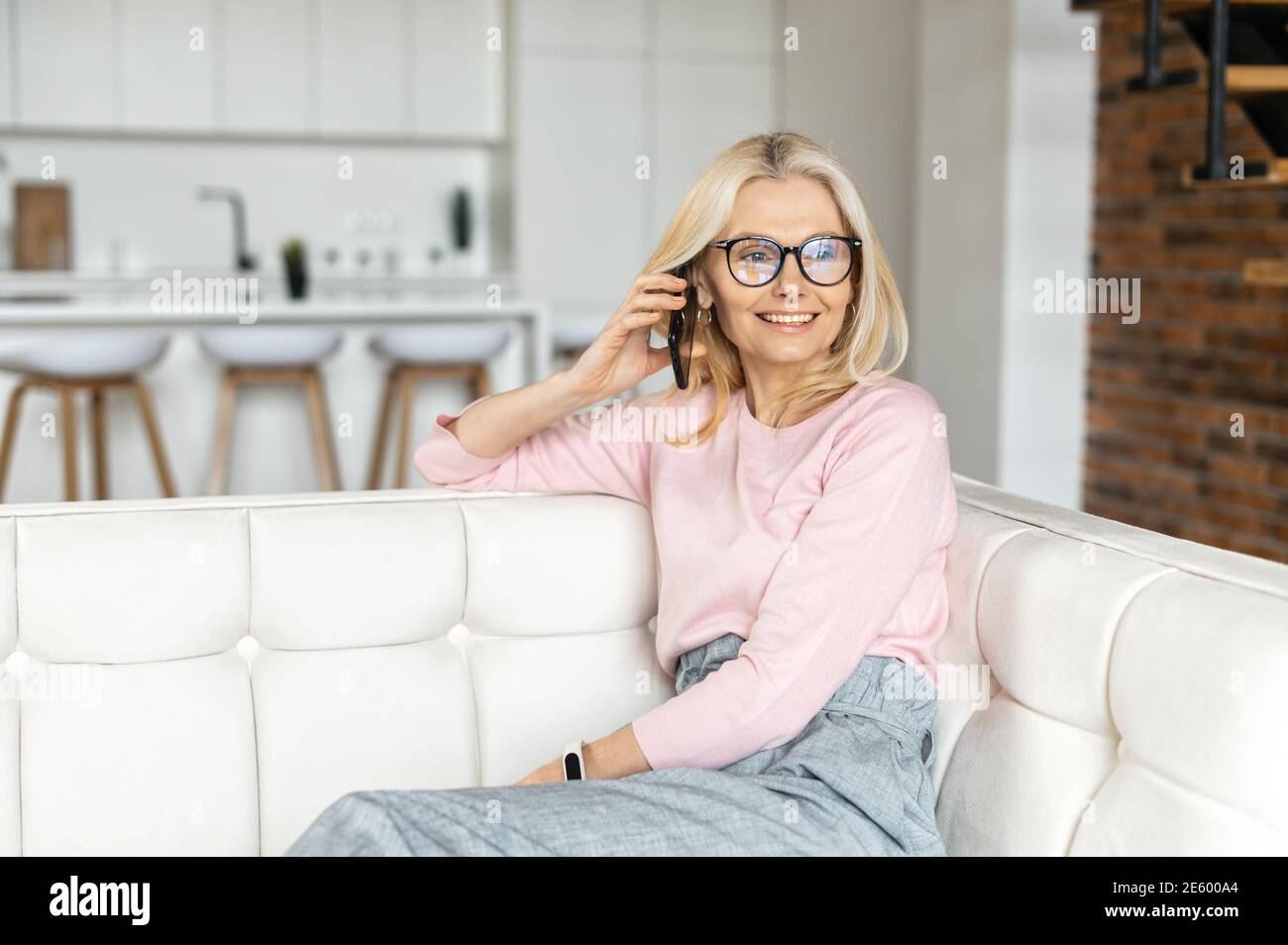 Elegante donna di mezza età indossando occhiali ha conversazione telefonica, una donna di affari rilassato si siede sul divano in appartamento moderno, ridere e. Foto Stock