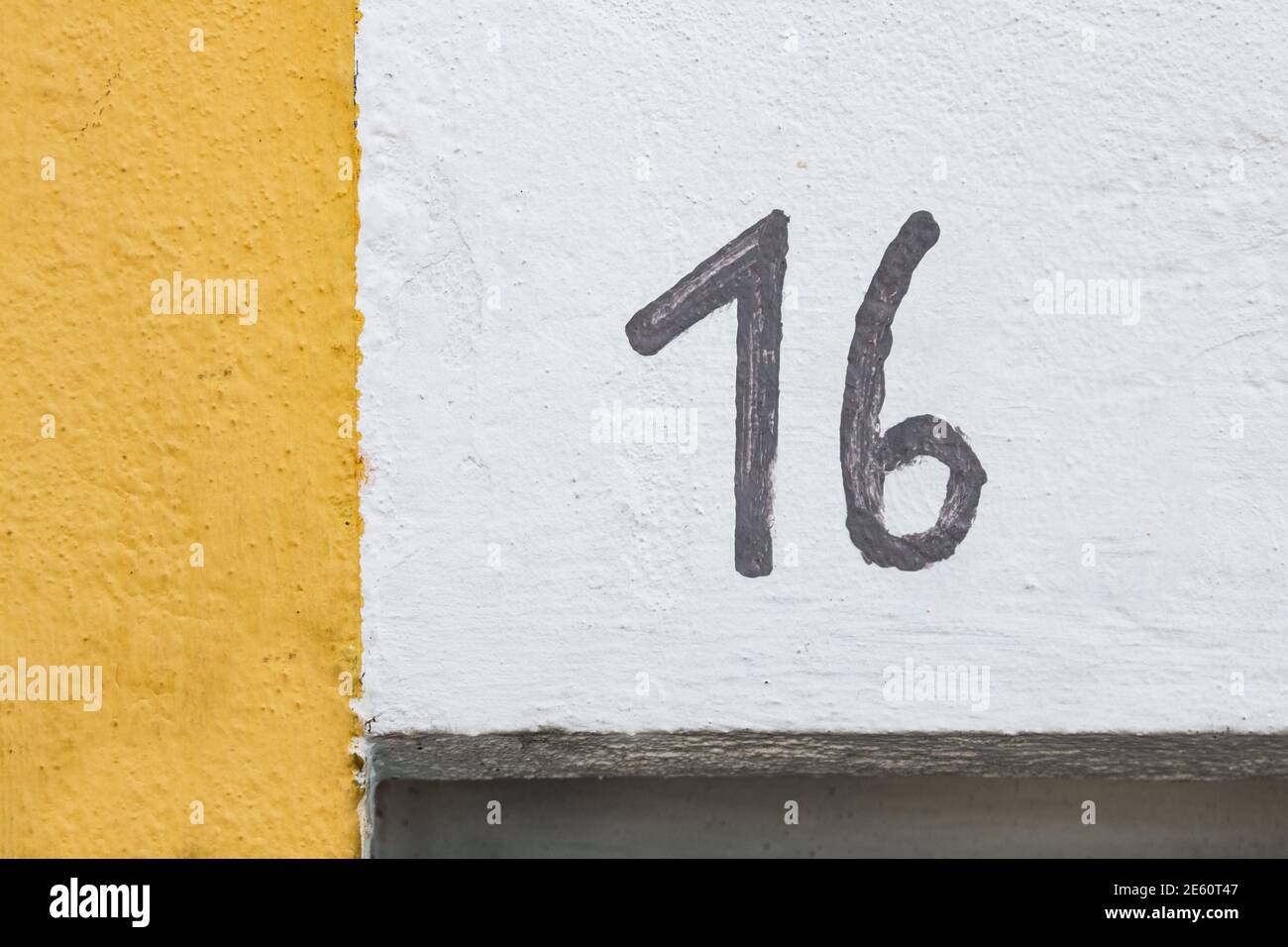 Il numero 16 è approssimativamente dipinto a mano in nero su una parete bianca con una striscia di inchiostro gialla sul lato sinistro del telaio. Particolare della facciata con numero civico Foto Stock Il numero 16 è approssimativamente dipinto a mano in nero su una parete bianca con una striscia di inchiostro gialla sul lato sinistro del telaio. Particolare della facciata con numero civico Foto Stock