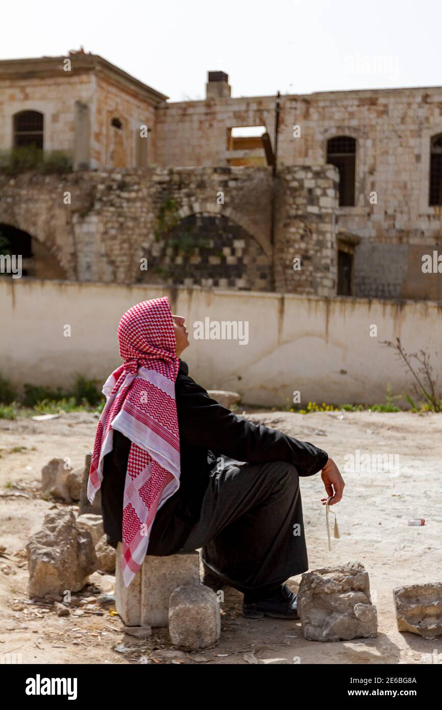 Hama, Siria 04-02-2010: Un giovane arabo che indossa Keffiyeh e abito da tobo jubba nero è seduto su una roccia nel quartiere della città vecchia di Hama e. Foto Stock