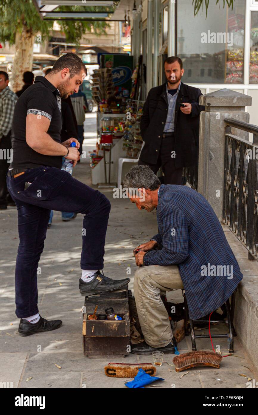 Hama, Siria 04-02-2010: Un anziano siriano Shiner Shoe sta lucidando le scarpe di un giovane uomo sulla sua scatola di shoeshine sul marciapiede da una strada affollata. Vecchio m Foto Stock