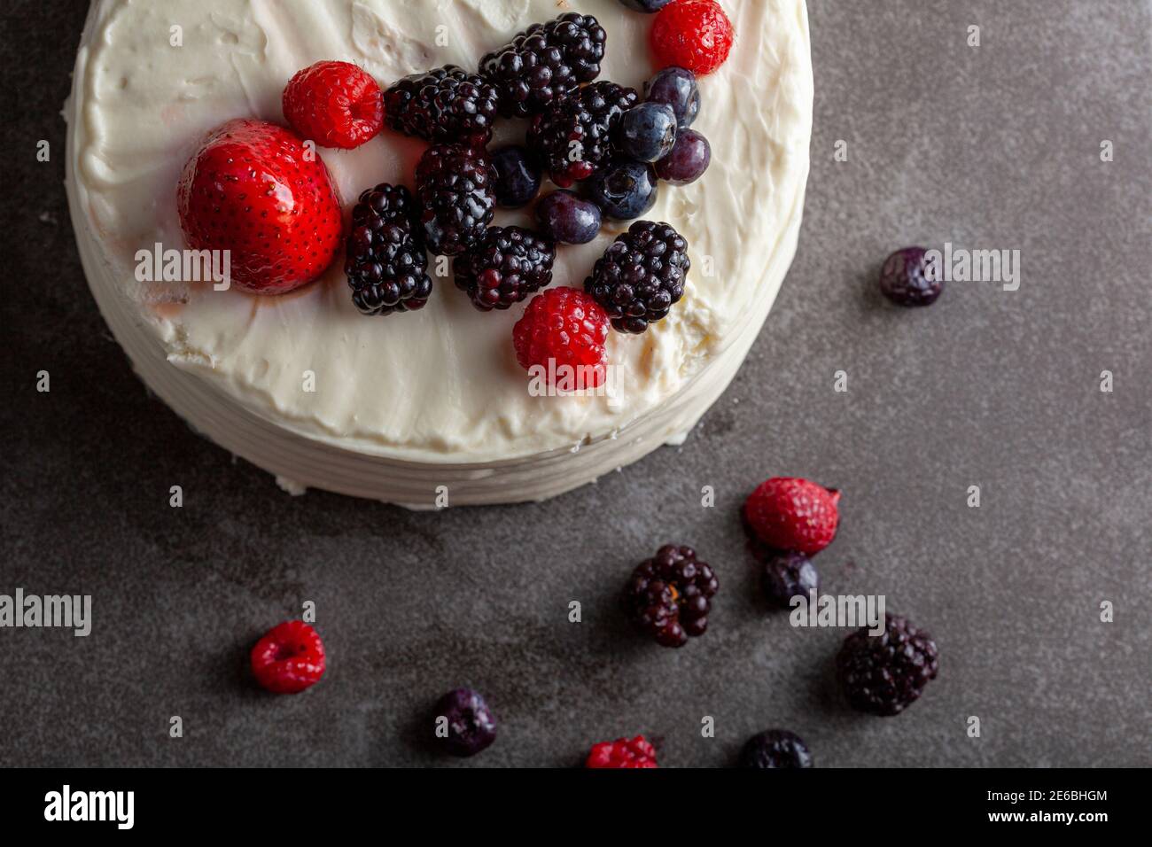 Primo piano immagine piatta di una torta rotonda con una glassa bianca cremosa e un assortimento di frutti di bosco utilizzati come guarnizioni decorative. Servito su grigio scuro b Foto Stock