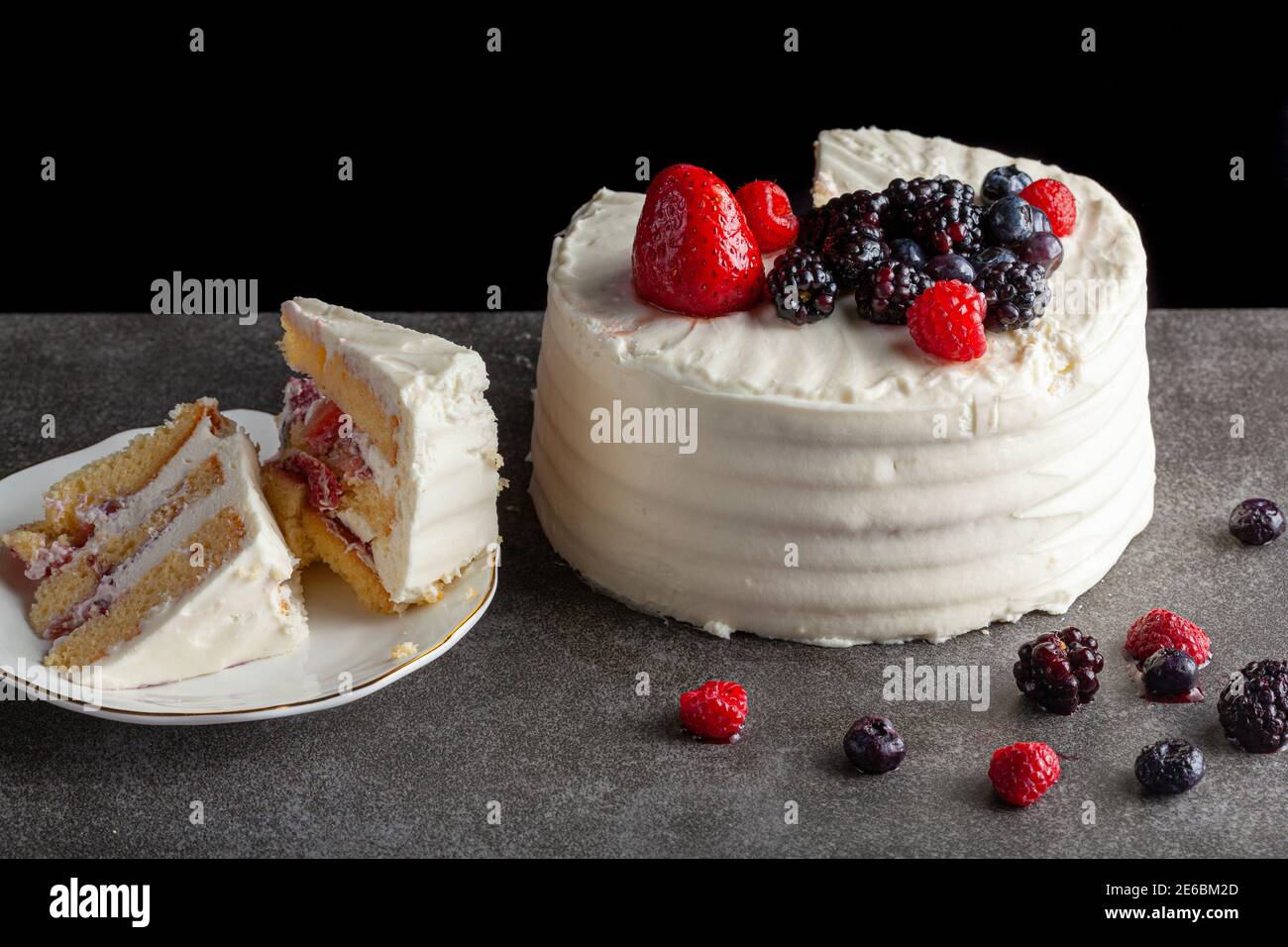 Primo piano immagine di una torta rotonda con glassa bianca cremosa e assortimento di frutti di bosco utilizzati come guarnizioni decorative. Servita su sfondo nero grigio scuro. TW Foto Stock