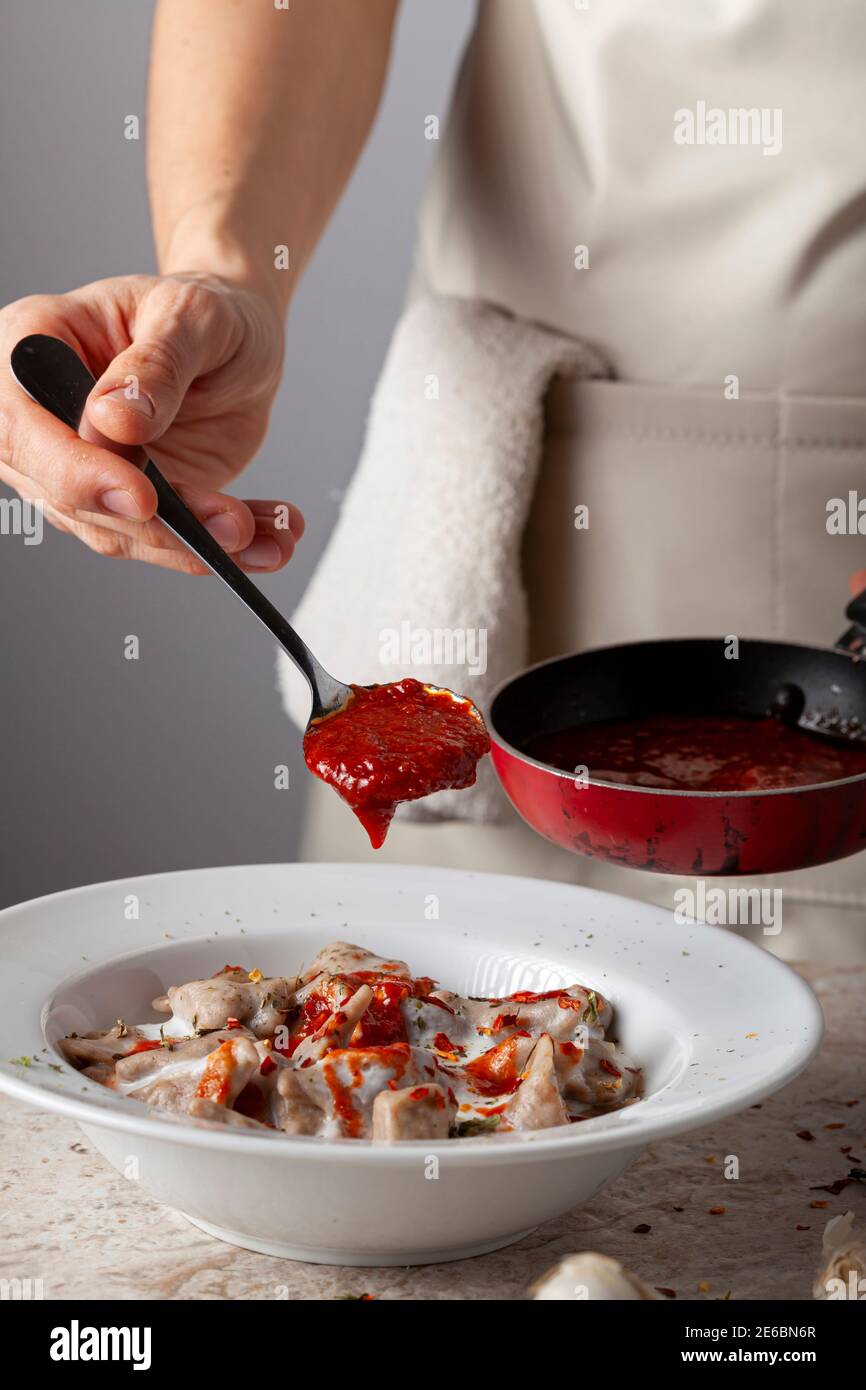 Una chef donna che indossa grembiule aggiunge salsa di pomodoro fresca ai gnocchi turchi fatti in casa nello yogurt. Questo piatto tradizionale è conosciuto come Manti ed è servito Foto Stock