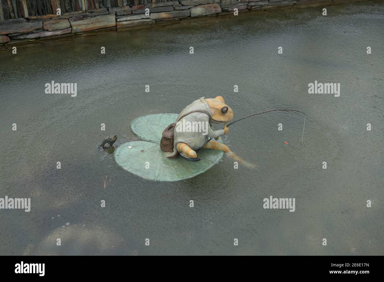 Rana di ceramica seduta su un giglio Pad con una canna da pesca in uno stagno congelato a Rosemoor Garden in Devon Rurale, Inghilterra, Regno Unito Foto Stock