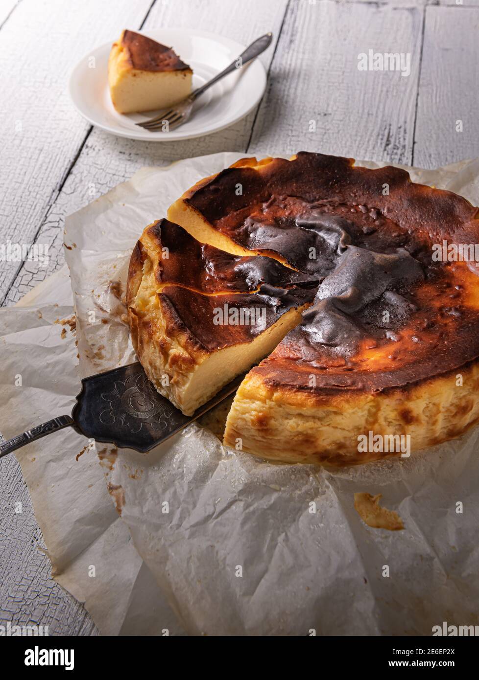 Primo piano della cheesecake basca tagliata bruciata sulla spatola di  servizio argentata in carta da forno su tavolo di legno bianco  testurizzato. Porzione non focalizzata sulla piastra sul retro Foto stock -