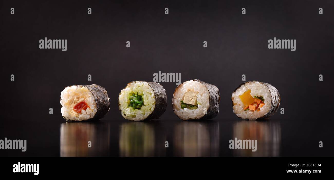 Assortimento di quattro sushi maki vegetali allineati su un tavolo di legno scuro con sfondo nero. Vista frontale. Composizione orizzontale. Foto Stock