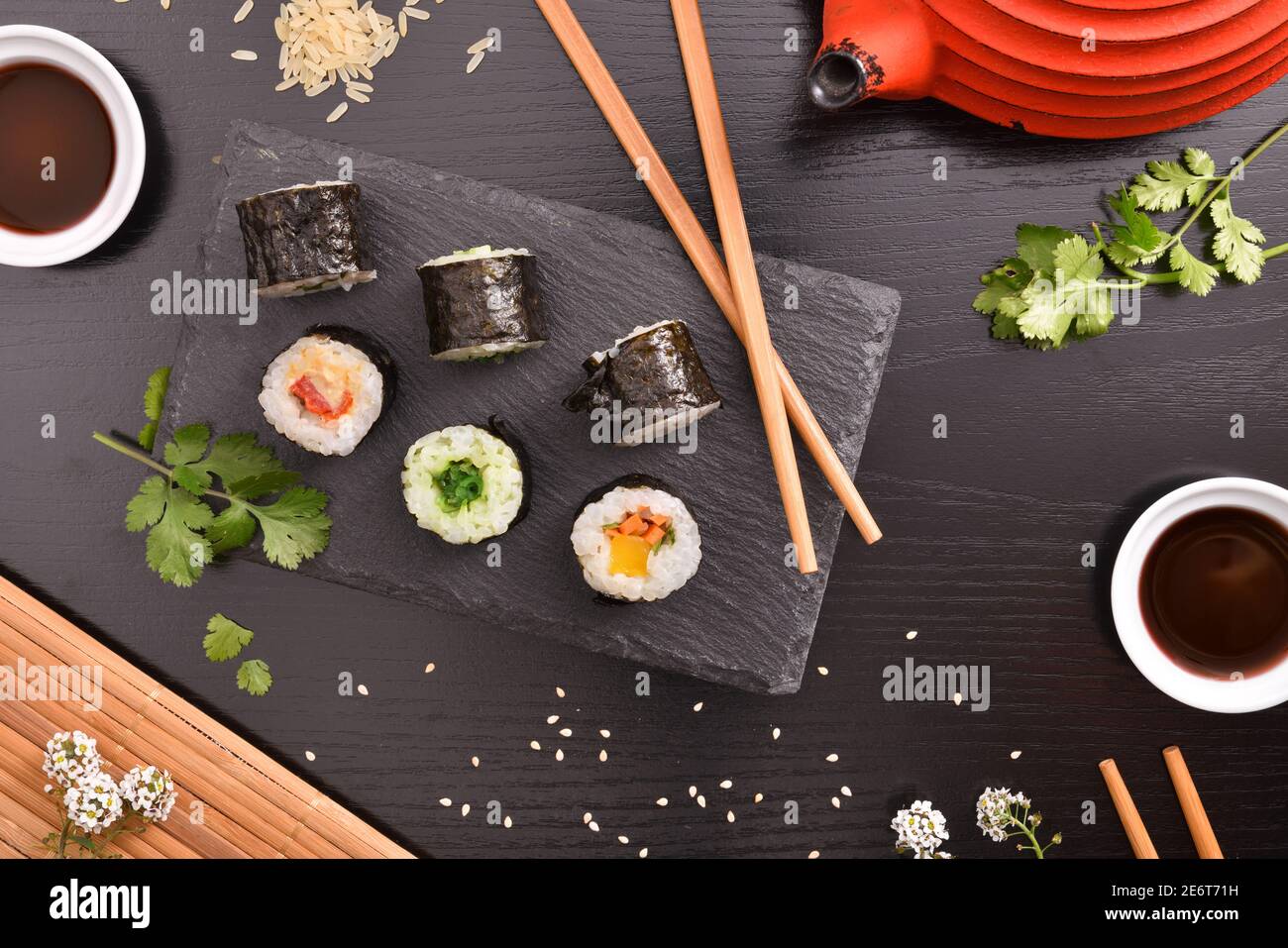 Sushi maki su piastra di ardesia e bastoncini a tavola nera con contenitori con salsa di soia, ingredienti e teiera. Vista dall'alto. Composizione orizzontale. Foto Stock