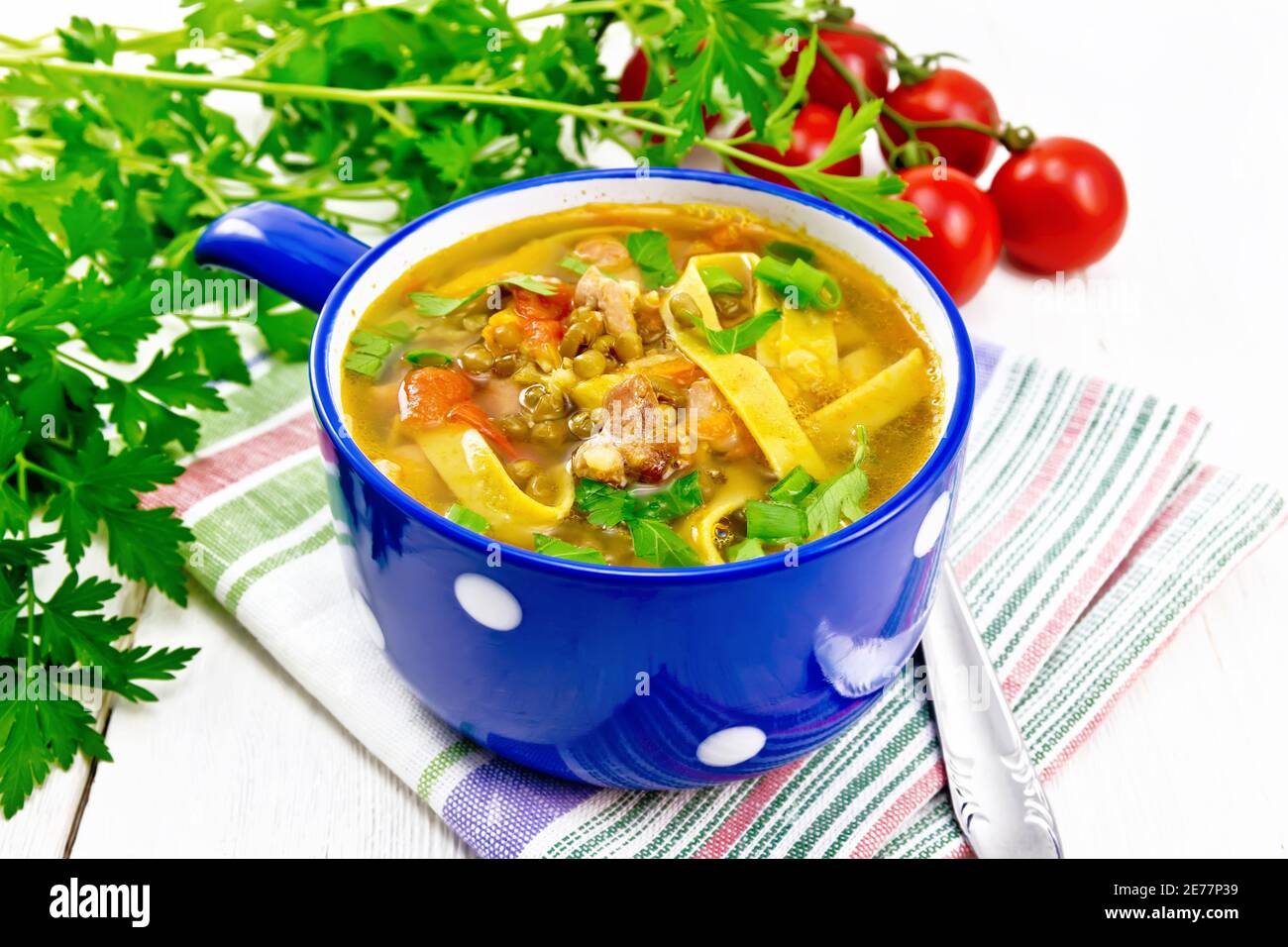 Zuppa con carne, pomodori, verdure, lenticchie di fagioli e tagliatelle in una ciotola blu su asciugamano, prezzemolo e un cucchiaio su sfondo di legno Foto Stock