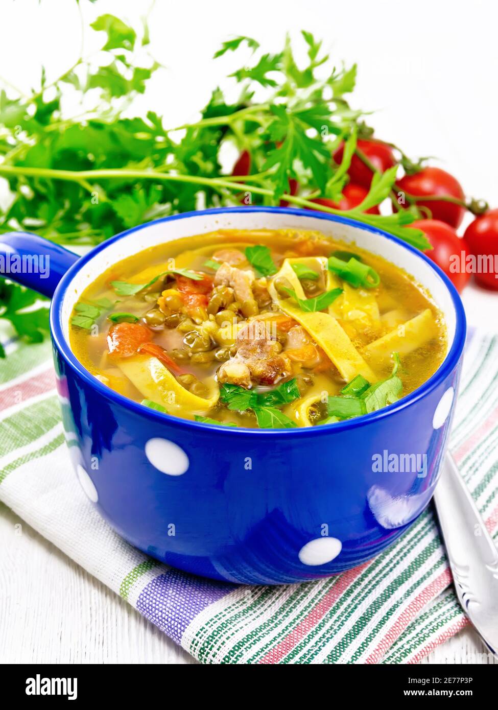 Zuppa con carne, pomodori, verdure, lenticchie di fagioli e tagliatelle in una ciotola blu su un tovagliolo, prezzemolo e cucchiaio su sfondo di legno Foto Stock