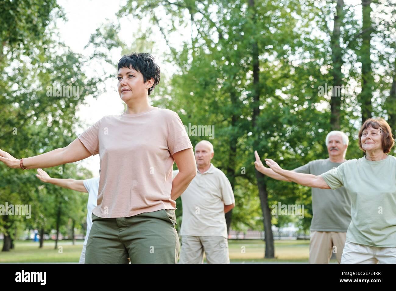 Donna matura allenarsi all'aperto nel parco con altri maturi persone sullo sfondo Foto Stock