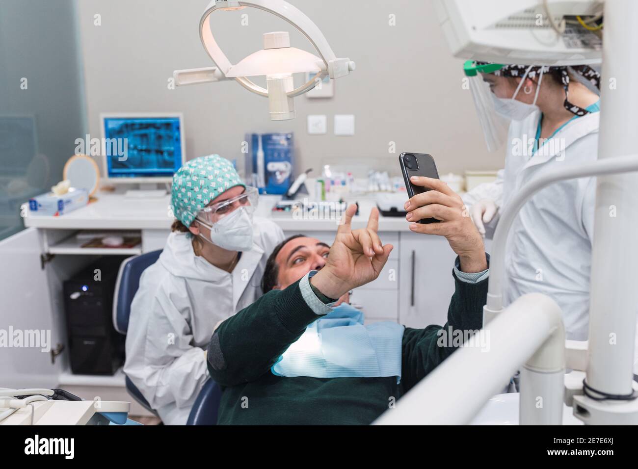 Paziente che prende un selfie nello studio dentistico. Tre persone. Medico femminile. Concetto di accoglienza e benessere. Sorrisi. Concentrarsi sullo smartphone. Foto Stock