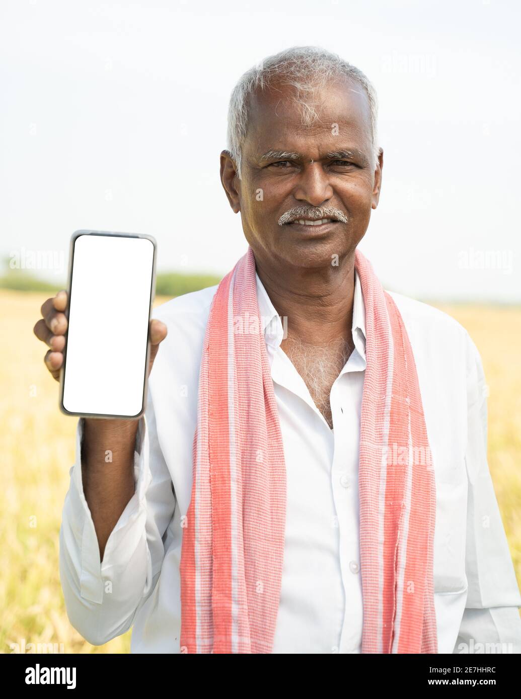 Sorridente vecchio agricoltore indiano in piedi di fronte a terreni agricoli vicino tenere il telefono cellulare mentre si guarda alla fotocamera Foto Stock