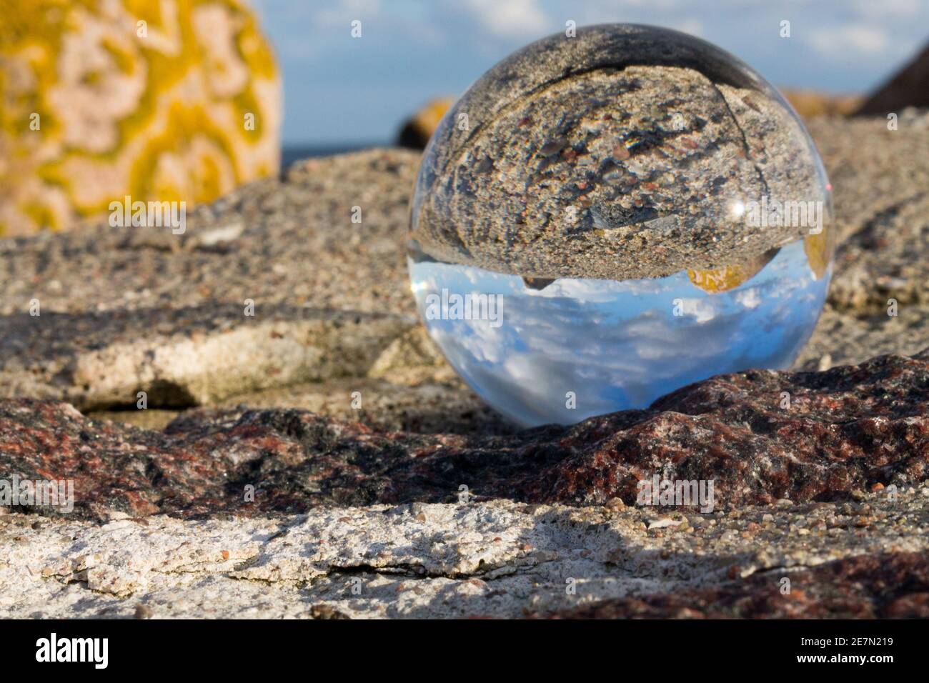Una sfera di cristallo poggia su una roccia ricoperta di licheni, che riflette un'immagine invertita del cielo blu e delle nuvole, mescolando texture naturali con meraviglia ottica. Foto Stock
