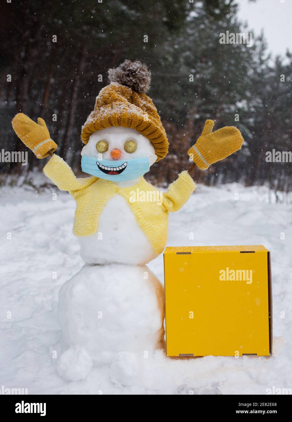 Felice pupazzo di neve in giallo maglia cappello, giacca, guanti e maschera  medica con sorriso dipinto su di esso. Nelle vicinanze si trova una scatola  di cartone giallo brillante con spazio per