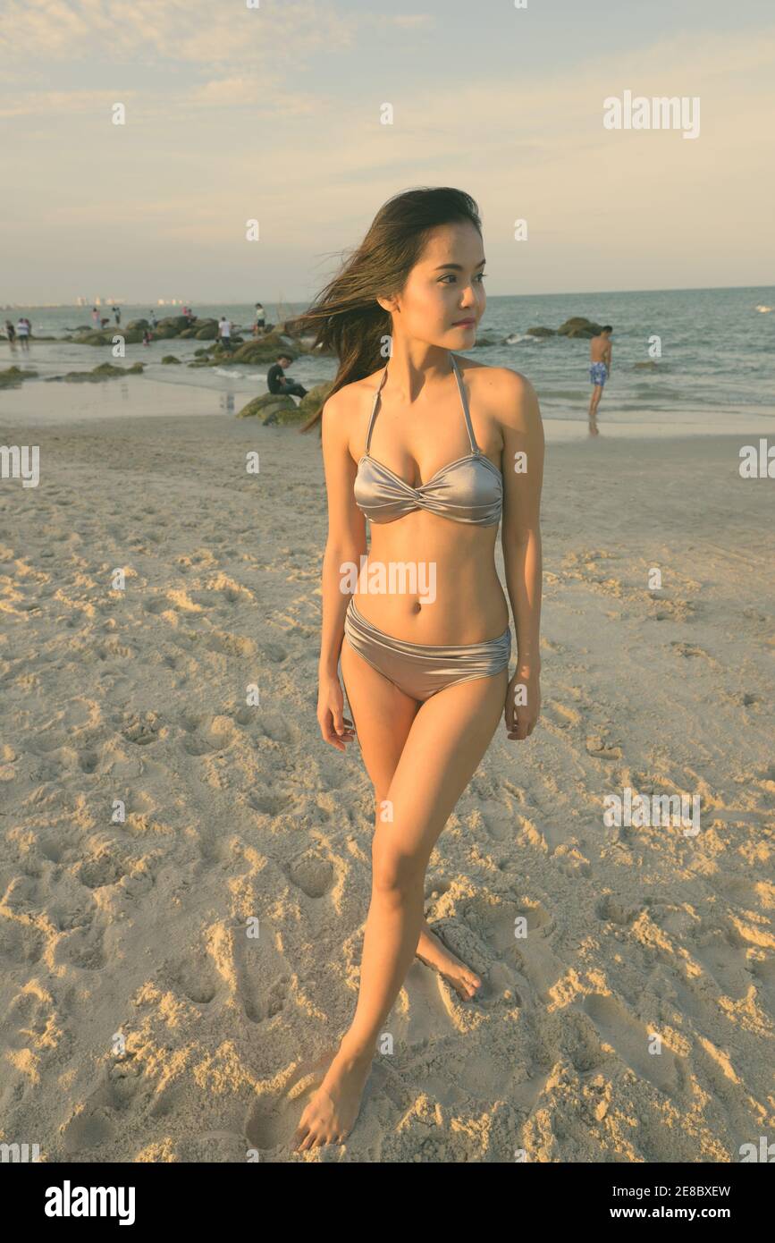 Giovane bella donna asiatica pensando mentre in piedi presso la spiaggia pubblica di Hua Hin in Thailandia con la luce soffusa del sole la trasmissione via IR Foto Stock