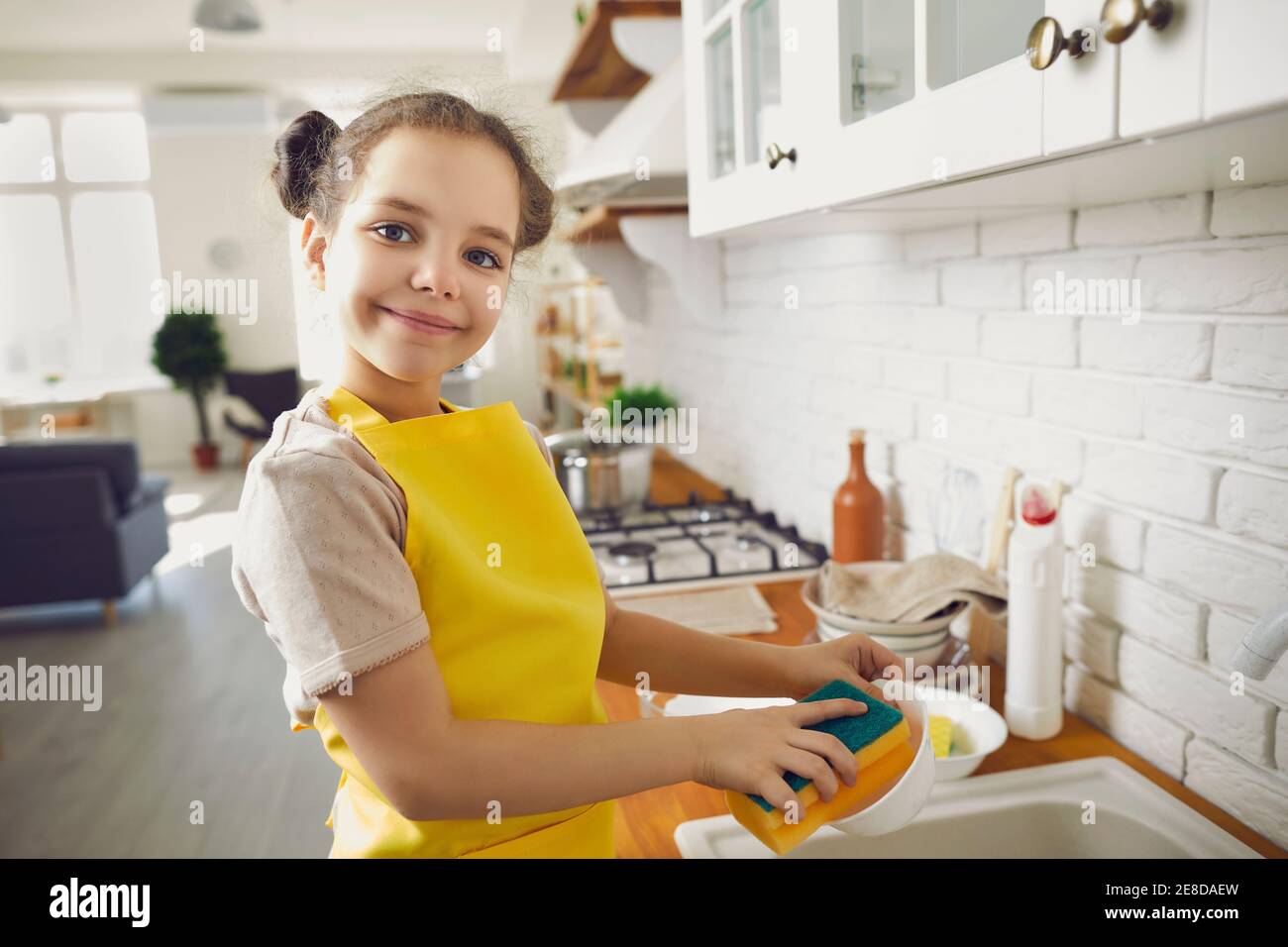 Ritratto di una bambina carina in un grembiule luminoso lavare i piatti in una cucina luminosa. Foto Stock