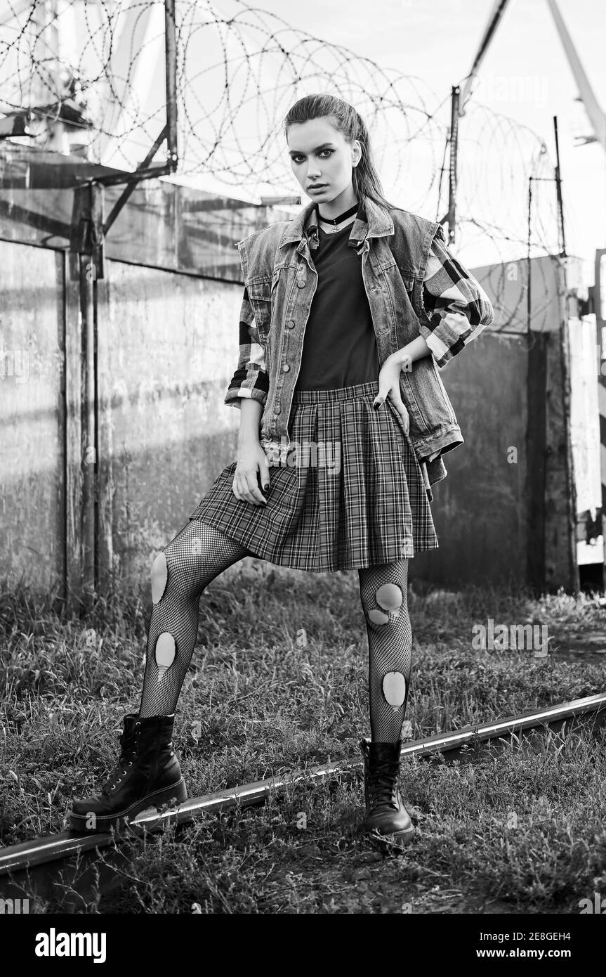 Ritratto esterno di ragazza grunge (rock) attraente in un luogo industriale. Modello informale vestito con giacca jean, gonna a scacchi e camicia, stivali e hol Foto Stock