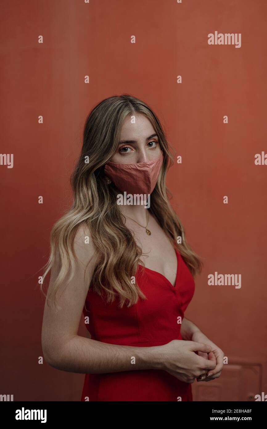 Anonimo giovane femmina dolce in vestito rosso e maschera in piedi con le mani aggrappate mentre si guarda la fotocamera durante la pandemia del coronavirus Foto Stock