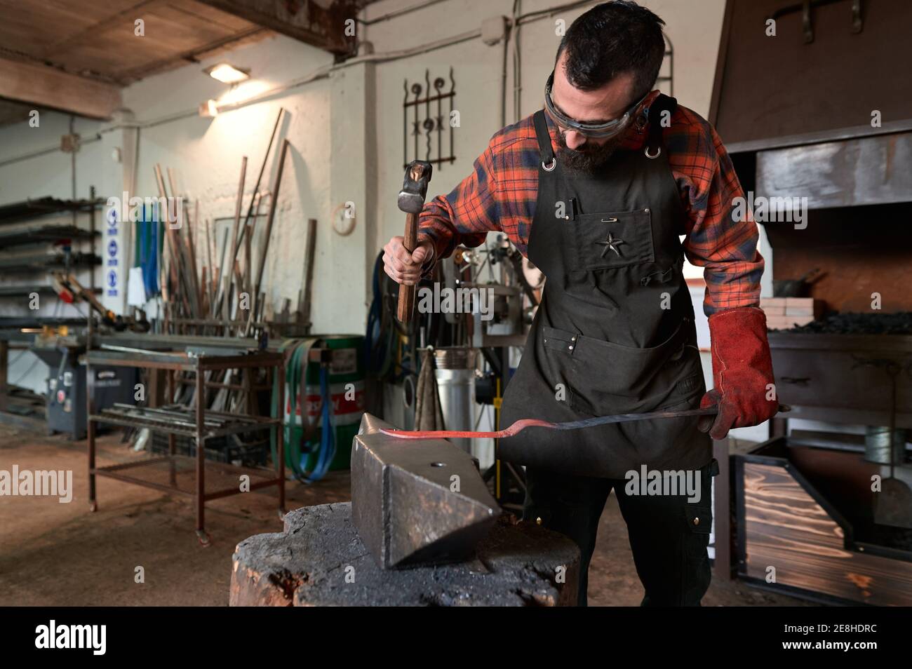 Concentrato giovane fabbro bearded in grembiule e occhiali di protezione colpisce metallo riscaldato con martello sull'incudine mentre si lavora in fucina Foto Stock