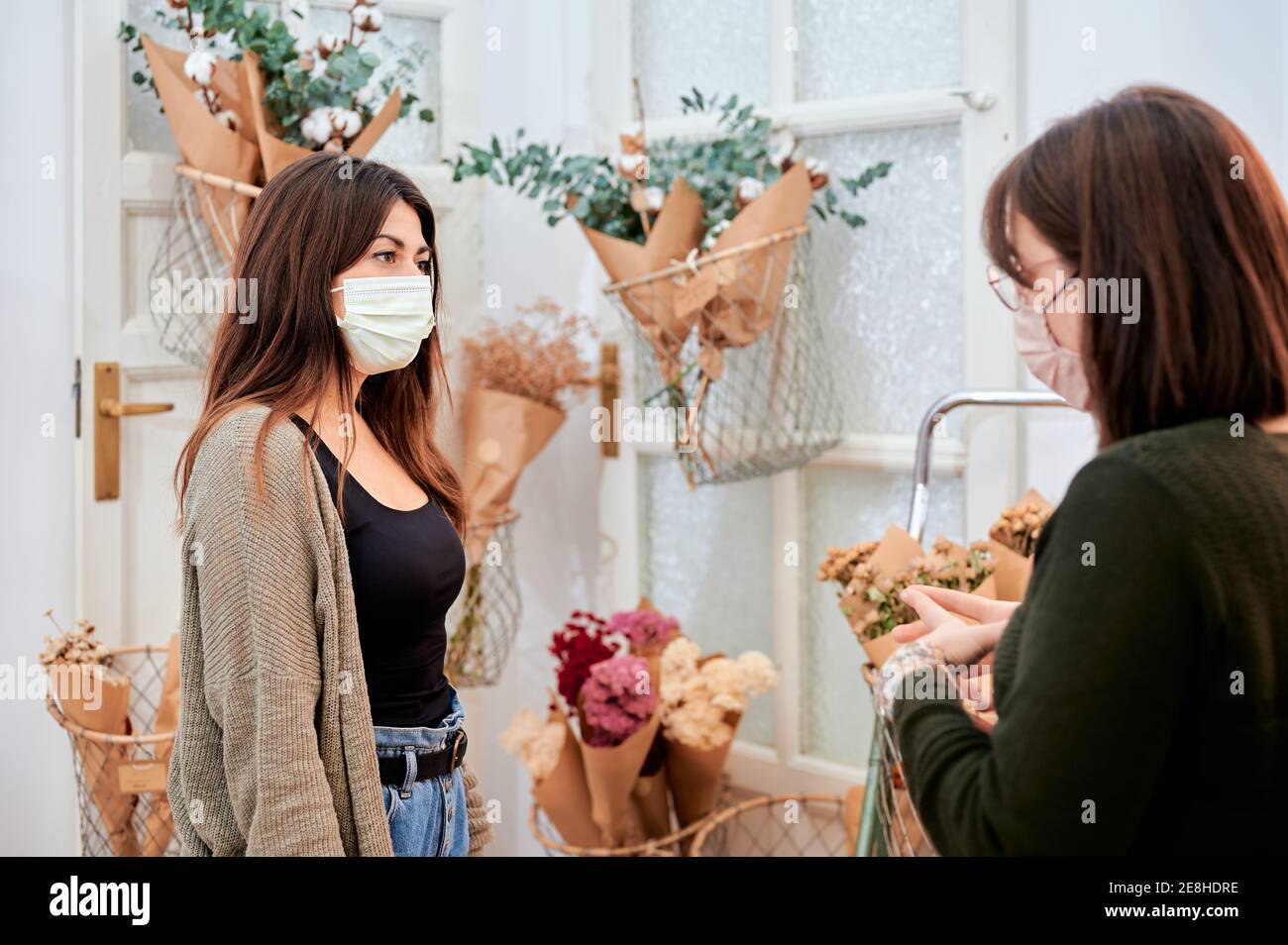 Cliente femminile in maschera protettiva consulenza con venditore che lavora in negozio con diversi mazzi di fiori avvolti in confezione di carta a zero rifiuti Foto Stock