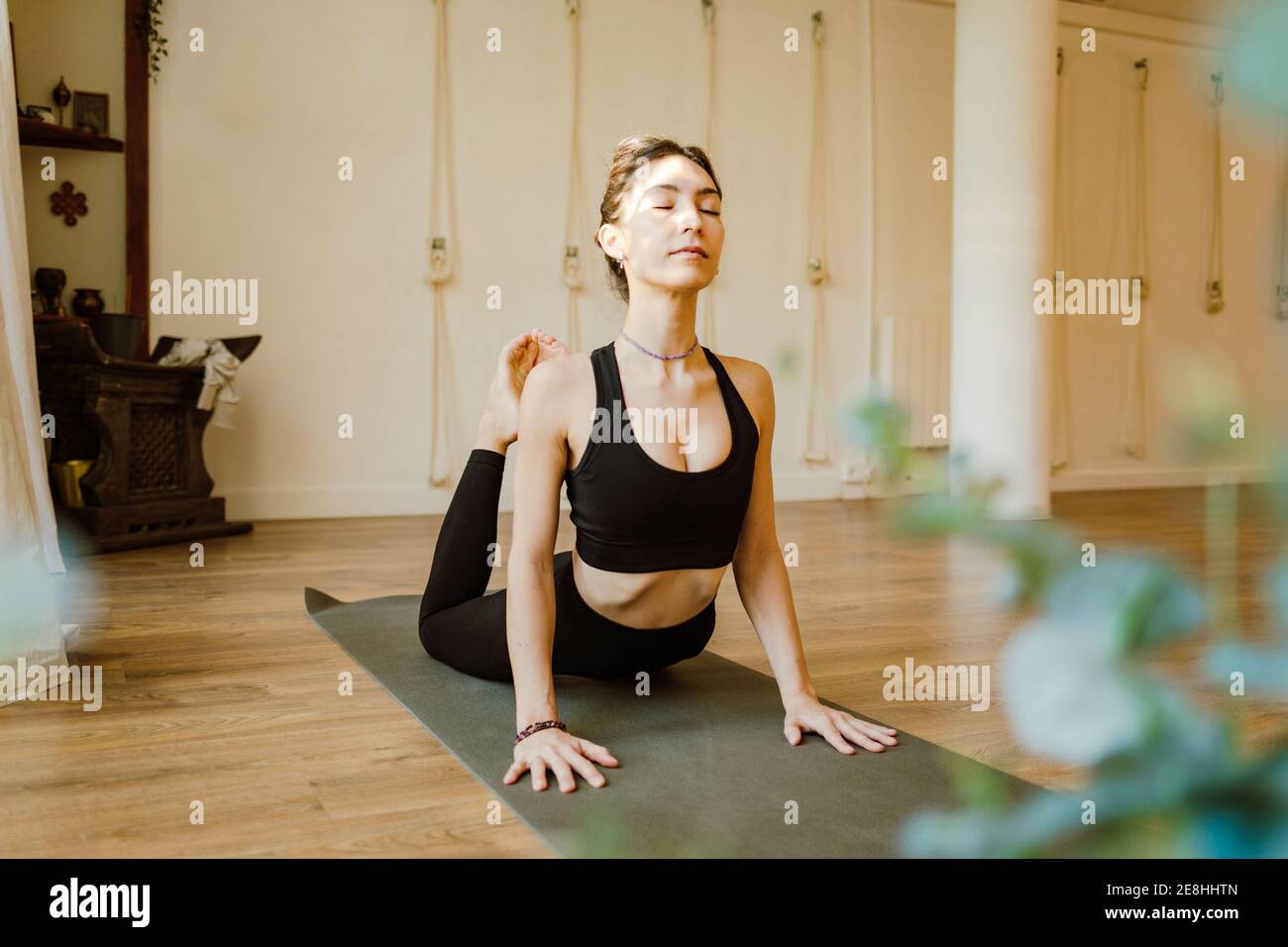 Giovane donna concentrata in abbigliamento sportivo che dimostra Raja Bhujangasana posa con occhi chiusi sul tappetino yoga in casa Foto Stock