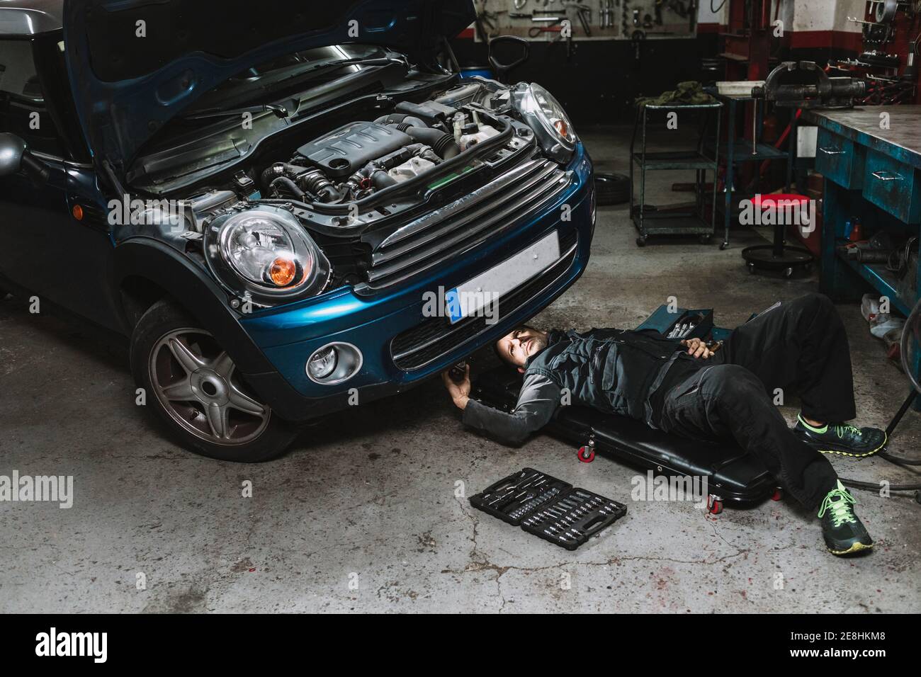 Giovane uomo di servizio auto focalizzato che controlla il veicolo con aperto cofano mentre si trova sul tappetino in officina Foto Stock